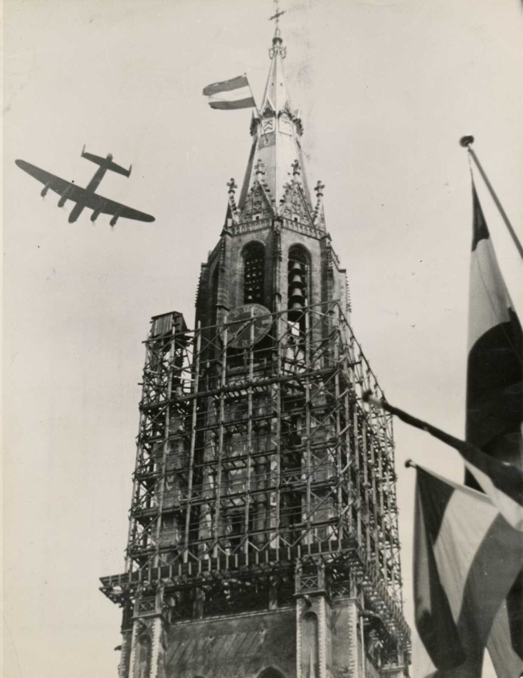 Een zwart wit foto van de Nieuwe Kerk na de bevrijding. Met stellages om de kerk en een vliegtuig in de lucht.